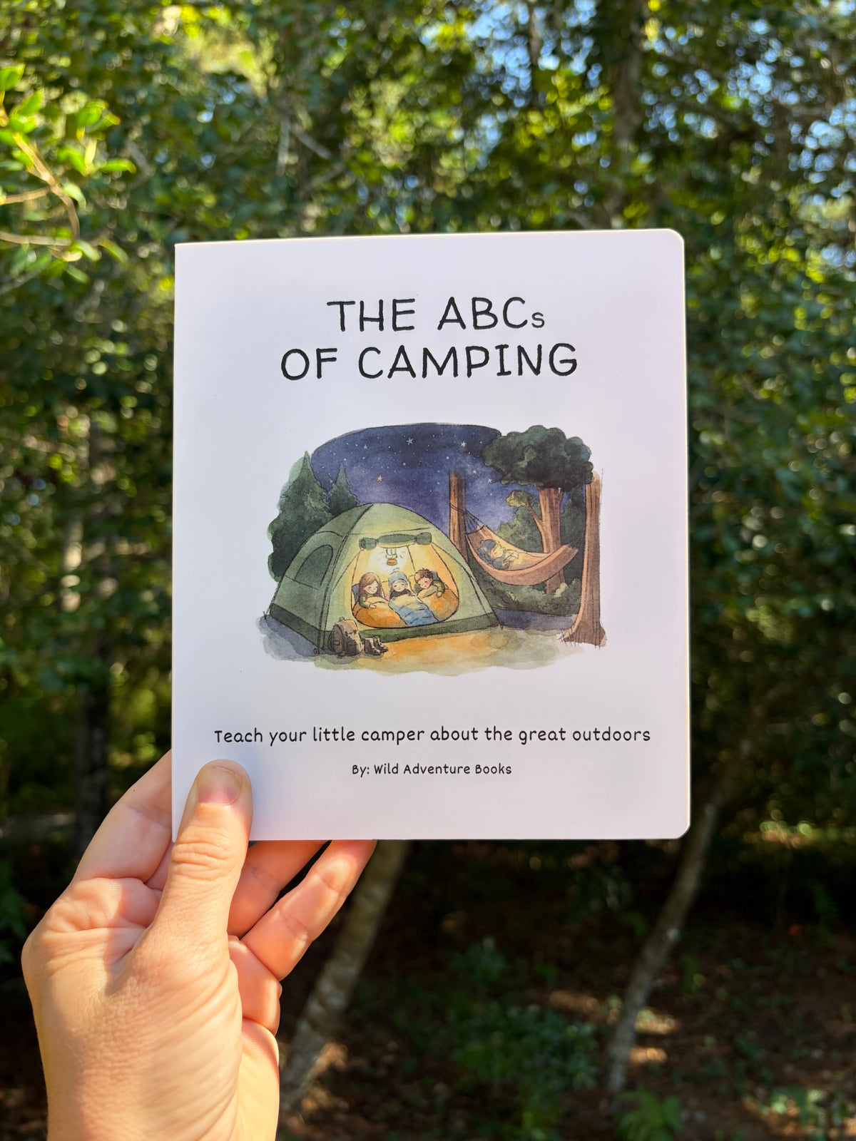A hand holds a children's book titled "The ABCs of Camping" against a blurred outdoor forest background. The book cover shows a camping scene with a tent, campfire, and hammock, and the text reads "Teach your little camper about the great outdoors" by Wild Adventure Books.
