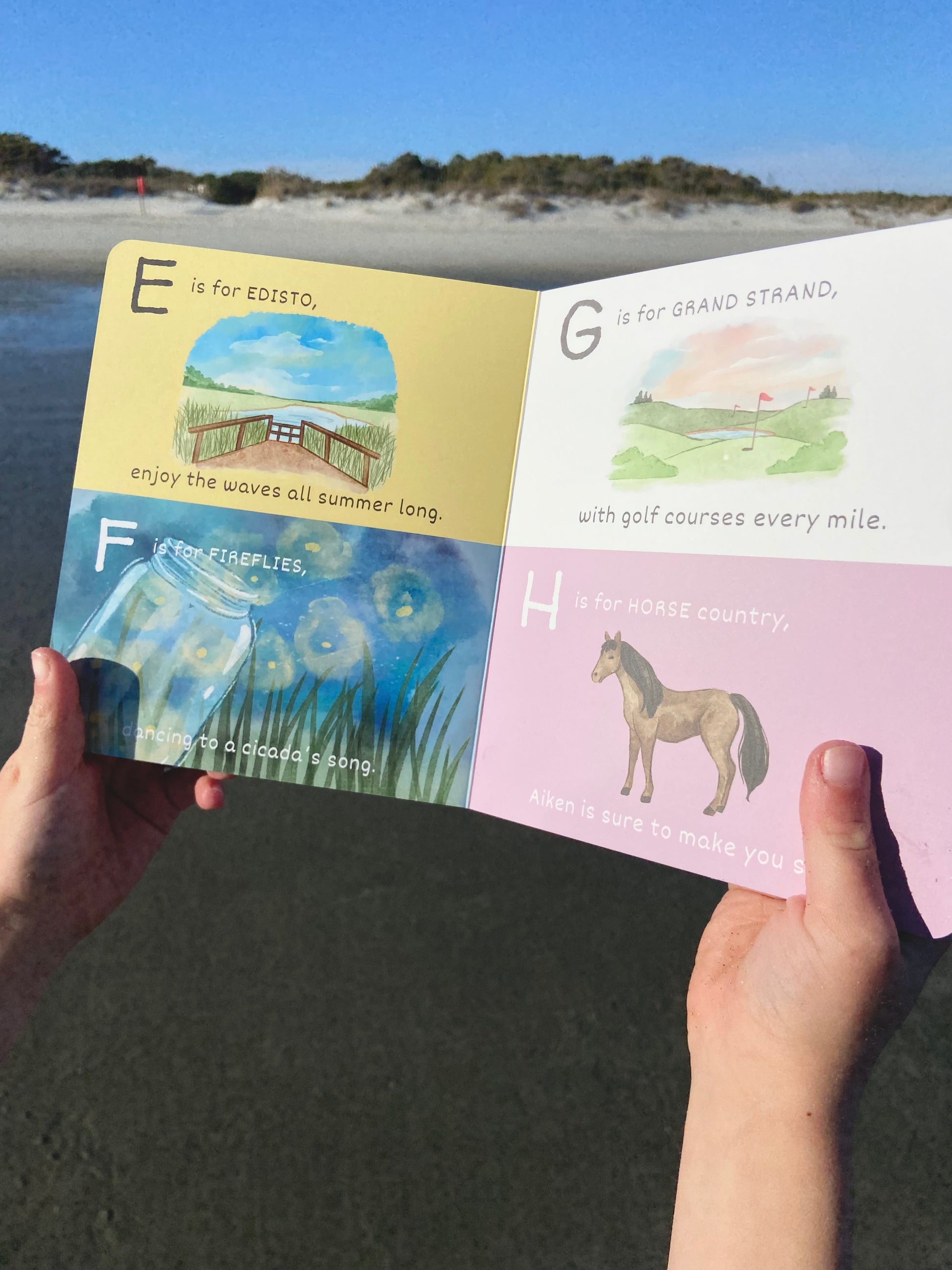Children's book open to pages with illustrations and text, held by hands against a beach background.